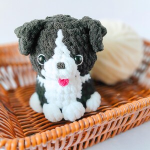 May include: A small, handmade crochet plush toy shaped like a puppy. The dog has dark charcoal gray fur with a white chest, face stripe, and paws. It features shiny black plastic eyes, a small brown nose, and a tiny bright pink yarn tongue.