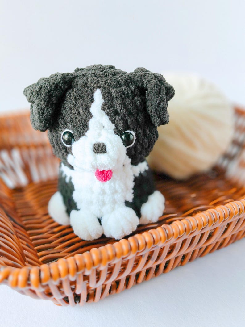 May include: A small, handmade crochet plush toy shaped like a puppy. The dog has dark charcoal gray fur with a white chest, face stripe, and paws. It features shiny black plastic eyes, a small brown nose, and a tiny bright pink yarn tongue.