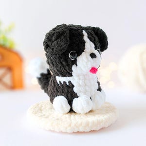 May include: A small, handmade crochet dog plushie featuring black and white yarn. The figure has a white face, chest, and paws, with black ears and body. It has two black plastic eyes and a small, bright pink yarn tongue protruding from its mouth.