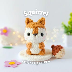 Puede incluir: Una figura de ardilla de ganchillo con una bellota a juego. La ardilla es blanca y marrón, con ojos negros y un pequeño rubor rosa. Se muestra el texto "Squirrel crochet pattern".