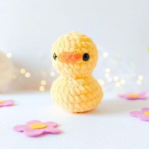 May include: A handmade, yellow crochet duckling with an orange beak and black eyes. The plush toy is surrounded by pink felt flowers with yellow centers. The background is white with soft, out-of-focus lights.