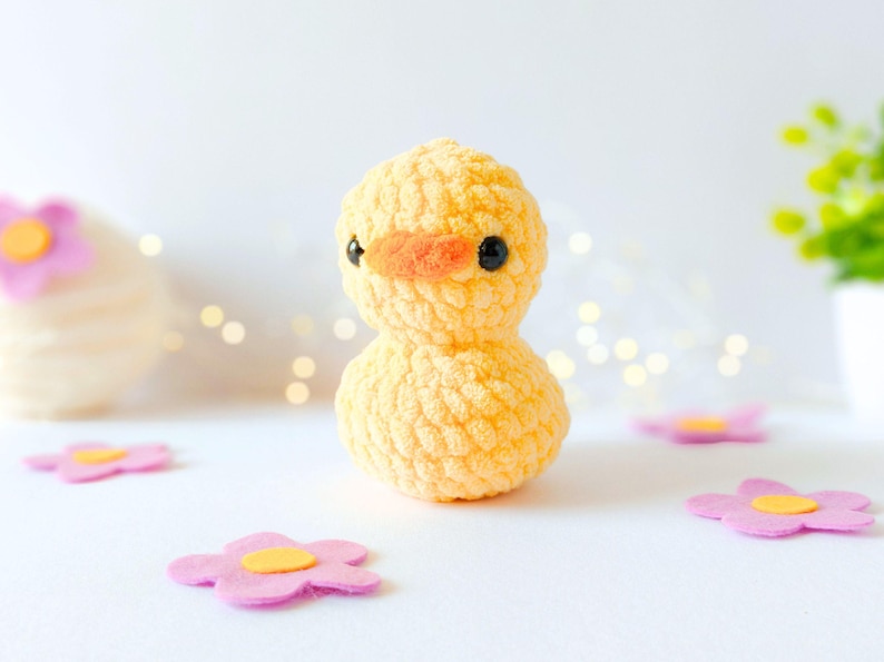 May include: A handmade, yellow crochet duckling with an orange beak and black eyes. The plush toy is surrounded by pink felt flowers with yellow centers. The background is white with soft, out-of-focus lights.