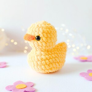 May include: A handmade yellow crochet duck with an orange beak and black eyes. The duck is sitting on a white surface, surrounded by pink felt flowers with yellow centers. The duck is a cute, whimsical toy.