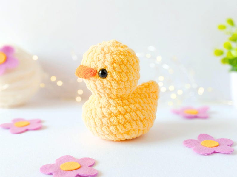 May include: A handmade yellow crochet duck with an orange beak and black eyes. The duck is sitting on a white surface, surrounded by pink felt flowers with yellow centers. The duck is a cute, whimsical toy.