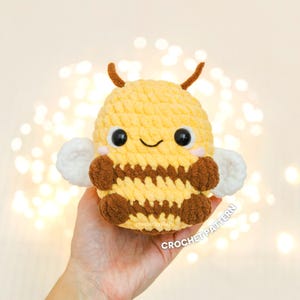 May include: A crocheted bee toy, predominantly yellow with brown stripes and details. It features white wings, brown antennae, and a smiling face. The text "CROCHET PATTERN" is visible on the bottom right.
