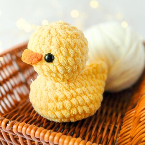 May include: A handmade yellow crochet duck with an orange beak and black eyes sits in a brown wicker basket. A ball of white yarn rests behind the duck. The duck is a soft, plush toy.