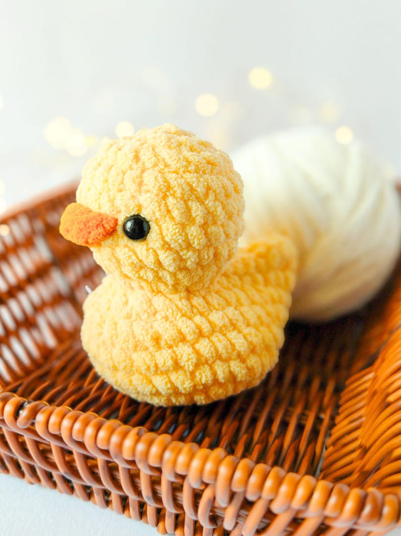 May include: A handmade yellow crochet duck with an orange beak and black eyes sits in a brown wicker basket. A ball of white yarn rests behind the duck. The duck is a soft, plush toy.
