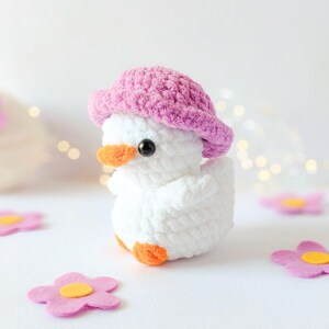 May include: A handmade white crochet duck wearing a pink hat with an orange beak and feet. The duck is surrounded by pink felt flower decorations. The background is white with soft lighting.