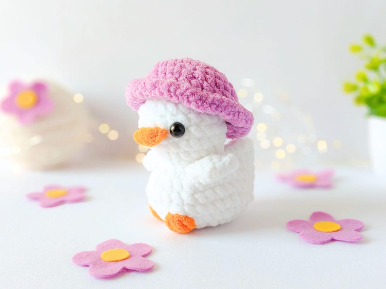 May include: A handmade white crochet duck wearing a pink hat with an orange beak and feet. The duck is surrounded by pink felt flower decorations. The background is white with soft lighting.