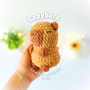 May include: A small, plush, brown capybara toy made with chunky crochet yarn. The toy features a darker brown snout and ears, with simple black stitched eyes and nose. The text "Capybara" and "crochet pattern no-sew" is printed in white above and below.