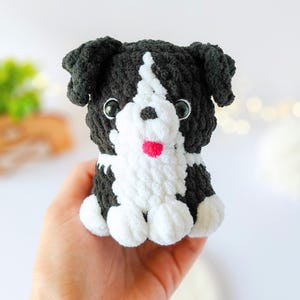 May include: A small, handmade crochet plush toy shaped like a puppy. The dog features black and white yarn, with a white chest and face stripe, black ears, two dark plastic eyes, a small black nose, and a tiny bright pink tongue.