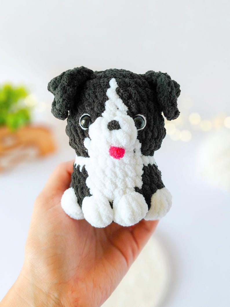 May include: A small, handmade crochet plush toy shaped like a puppy. The dog features black and white yarn, with a white chest and face stripe, black ears, two dark plastic eyes, a small black nose, and a tiny bright pink tongue.