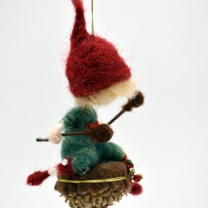 Puede incluir: Un adorno de gnomo de fieltro con un gorro rojo, un atuendo verde y una barba marrón. El gnomo está sentado en una bellota marrón con ribete dorado y cuentas rojas y doradas.