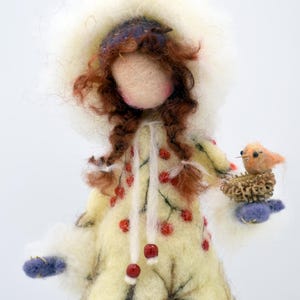 Könnte beinhalten: Eine Filzpuppe, die einen weißen Kapuzenmantel mit roten Beeren und einen blauen Hut trägt. Die Puppe hat langes braunes Haar und hält einen kleinen Vogel in einem Nest.