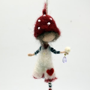 May include: A whimsical felted ornament of a mushroom-capped figure. The figure wears a white dress with a red heart, striped socks, and red shoes. It holds a small white puff and a purple teardrop-shaped charm. The mushroom cap is red with white spots.