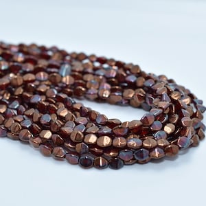 May include: A strand of small, metallic copper-colored beads with a smooth, irregular, flattened shape. The beads feature a reflective, iridescent finish that catches the light, creating subtle shifts in tone across the curved, polished surfaces.