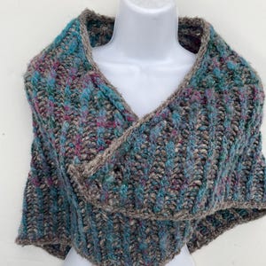May include: A hand-knitted shawl in shades of teal, gray, and magenta, displayed on a mannequin. The shawl features a textured, ribbed pattern with a contrasting gray border. The chunky knit design suggests warmth and comfort, perfect for cooler weather.