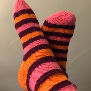 May include: A pair of knitted socks with horizontal stripes in shades of pink, orange, and burgundy. The socks are visible from the ankles to the toes, showcasing the colorful pattern. The socks appear to be handmade.