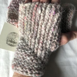May include: Hand-knitted fingerless gloves in shades of grey, white, and pink. The chunky knit features flecks of colour throughout. The gloves are designed to cover the hand and wrist, leaving the fingers free. A small tag is attached.
