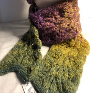 May include: A handmade knitted scarf and hat set. The scarf is a mix of purple, yellow, and green yarn, with a textured pattern. The hat is visible at the top of the image. The scarf is draped over a white mannequin head.