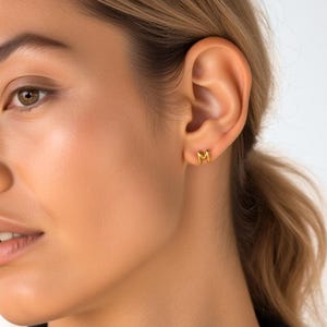 Może przedstawiać: Zbliżenie osoby noszącej złote kolczyki z literą "M". Kolczyki są małe i proste, z polerowanym wykończeniem. Osoba ma jasną skórę i ma na sobie ciemnoniebieski top.