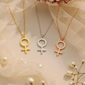 May include: Three necklaces with a female symbol pendant. One is gold, one is silver, and one is rose gold. The pendants are connected to delicate chains. The background is a soft, neutral color with scattered pearls.