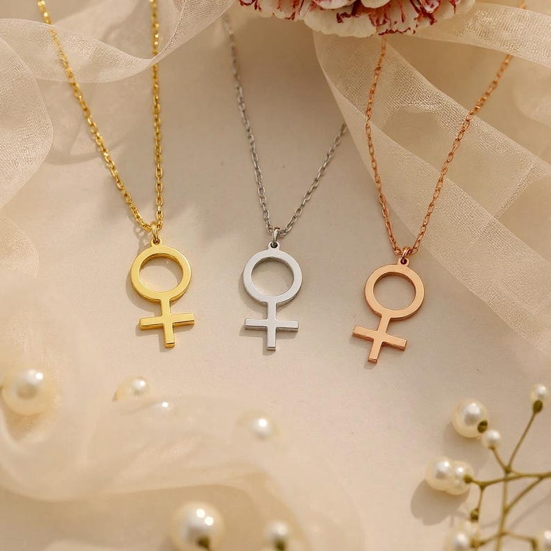 May include: Three necklaces with a female symbol pendant. One is gold, one is silver, and one is rose gold. The pendants are connected to delicate chains. The background is a soft, neutral color with scattered pearls.