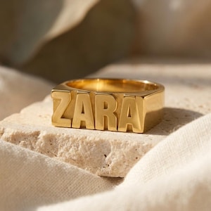 Può includere: Un anello a sigillo in metallo color oro con il nome ZARA in lettere maiuscole spesse e in rilievo sulla parte anteriore. L'anello presenta una superficie lucida e riflettente con un design a fascia larga e piatta posizionato orizzontalmente.