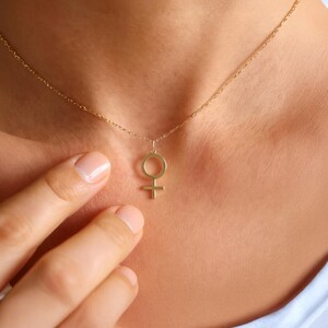 May include: A gold-colored necklace featuring a pendant in the shape of the Venus symbol. The pendant is suspended from a delicate chain. The necklace is worn around the neck.