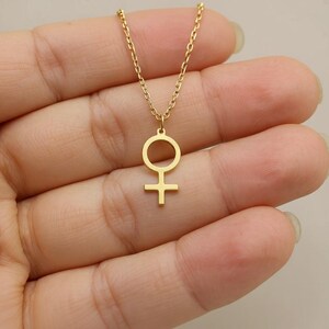 May include: A gold-colored necklace featuring a pendant with the Venus symbol. The pendant is a circle with a cross below it, suspended from a delicate chain. The necklace is a simple, elegant piece of jewelry.