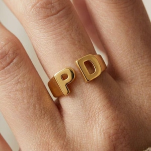May include: A gold-toned open ring featuring two prominent, raised capital letters, P and D, positioned at the ends of the band. The letters have a smooth, polished finish and are integrated into the structure of the ring, creating a bold, letter-based design.