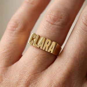 May include: A gold-toned metal ring featuring the name CLARA in raised, block-style capital letters across the front band. The ring has a polished, reflective surface and a smooth, solid band that wraps around the finger for a secure fit.