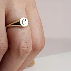May include: A gold signet ring with a rounded rectangular face. The face of the ring features the letter "C" in a delicate, serif font. The ring is worn on a finger, with the band tapering towards the back.