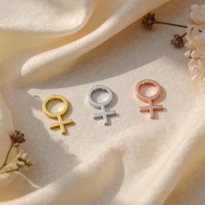 May include: Three metal pendants in gold, silver, and rose gold, each shaped as the Venus symbol. The pendants are displayed on a cream-colored, textured fabric, with small floral accents in the background. The pendants are small and delicate.