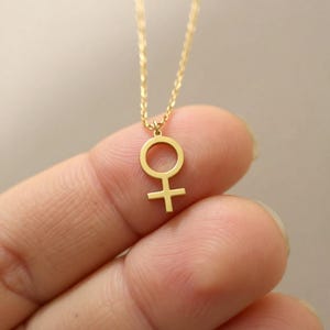 May include: A delicate gold-colored necklace featuring a pendant in the shape of the female symbol. The pendant is suspended from a fine chain. The necklace is shown against a blurred background.