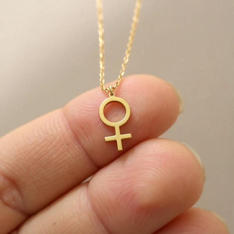 May include: A delicate gold-colored necklace featuring a pendant in the shape of the female symbol. The pendant is suspended from a fine chain. The necklace is shown against a blurred background.