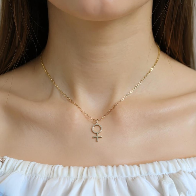May include: A gold-colored necklace with a pendant featuring the Venus symbol, a circle with a cross below it. The necklace is worn around the neck, and the pendant hangs at the chest. The chain is delicate and the pendant is small.