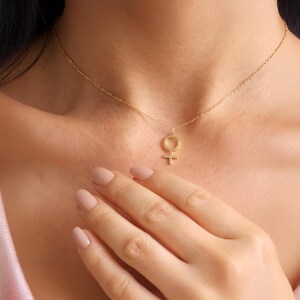 May include: A gold-colored necklace featuring a pendant in the shape of the female gender symbol. The delicate chain is draped around a person's neck, with the pendant resting near the collarbone. The person's hand is visible, with painted nails.