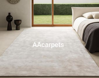May include: A large, rectangular, light gray area rug with a soft, uniform texture covering the floor. The rug features the text AAcarpets printed in white in the center. The rug is positioned in a room with a bed and decorative vases.