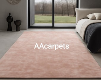 Op de afbeelding: Een rechthoekig vloerkleed in een effen, gedempte oudroze kleur met de witte tekst AAcarpets in het midden. Het kleed ligt op een gladde, lichtgrijze vloer in een kamer met een bed en donkere decoratieve vazen.