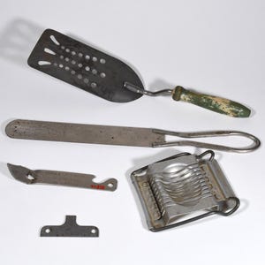 May include: A selection of antique kitchen tools, including a slotted spatula with a green and white handle, a long metal knife, a can opener, an egg slicer, and a small metal bracket. The items are displayed on a white background.