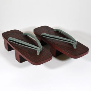 May include: Pair of traditional Japanese geta sandals. The wooden platform sandals are dark reddish-brown with a textured surface. The thong straps are light green with a black stripe. The sandals are elevated by two rectangular blocks.