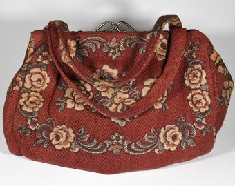 Puede incluir: Bolso de mano de estilo vintage en un tono marrón rojizo, adornado con patrones florales en beige y marrón. El bolso presenta una forma estructurada, un cierre metálico y un asa a juego. El diseño floral se muestra prominentemente en la parte delantera y los laterales.