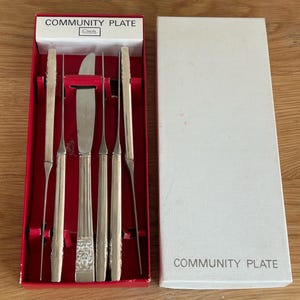 May include: A Community Plate silverware set in a red-lined box with a white lid. The set includes five silver-toned utensils with ornate handles, featuring a central knife with a floral pattern and four slender, pointed implements.