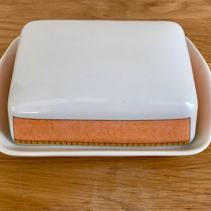 May include: A rectangular ceramic butter dish with a white lid and a matching white base. The lid features a wide, textured peach-coloured horizontal band around the bottom edge, accented by a thin, dark-outlined yellow grid pattern at the base.