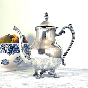 May include: A polished silver teapot with a curved spout and ornate handle. The teapot has a decorative lid and sits on three small feet. A blue and white patterned bowl with fruit is in the background.