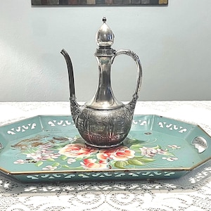 May include: A silver-colored, ornate pitcher with a long spout and handle sits on a turquoise, octagonal tray. The tray features a floral design with red and pink roses and green leaves. The tray has a gold trim.