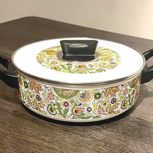 May include: A vintage cooking pot with a white lid and a floral pattern in shades of green, orange, and yellow. The pot has black handles and a black knob on the lid. The pot is sitting on a dark wooden surface.