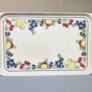 May include: A rectangular off-white serving tray featuring a decorative border of colorful fruit illustrations, including red cherries, yellow pears, purple plums, and dark blue grapes with green leaves, all set against a plain cream-colored center.