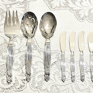 May include: A set of silver serving utensils, including a fork, slotted spoon, solid spoon, and four butter knives. The handles have a ribbed design with decorative detailing at the base. The silverware is arranged on a white, patterned tablecloth.